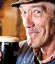 Een Ierse man met een hoed en een kleurrijke sjaal lacht terwijl hij geniet van een pint Guinness in een pub, de essentie van vakantie Ierland vastleggend. De achtergrond onthult wazige planken vol met drankjes, die de warme gastvrijheid van sfeervolle hotels belichamen.