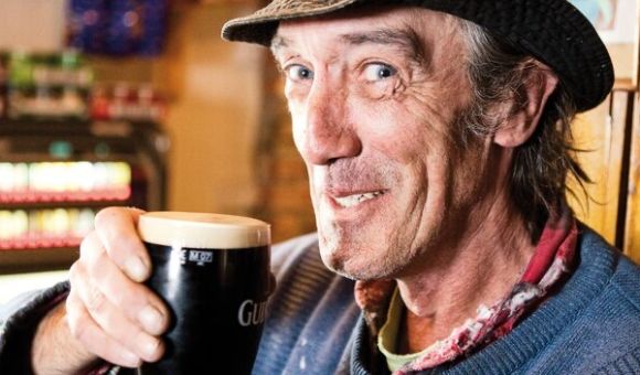 Een Ierse man met een hoed en een kleurrijke sjaal lacht terwijl hij geniet van een pint Guinness in een pub, de essentie van vakantie Ierland vastleggend. De achtergrond onthult wazige planken vol met drankjes, die de warme gastvrijheid van sfeervolle hotels belichamen.