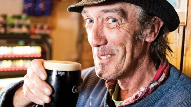 Een Ierse man met een hoed en een kleurrijke sjaal lacht terwijl hij geniet van een pint Guinness in een pub, de essentie van vakantie Ierland vastleggend. De achtergrond onthult wazige planken vol met drankjes, die de warme gastvrijheid van sfeervolle hotels belichamen.