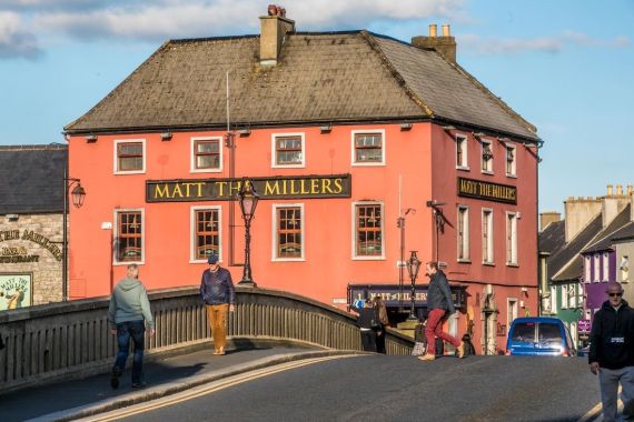 Een levendig, zalmkleurig gebouw met een bord met de tekst "Matt The Millers" staat op een straathoek en legt de charme van een rondreis Ierland vast. Meerdere mensen lopen over een gebogen brug die naar de pub leidt. De heldere lucht en de naderende kleine blauwe auto maken dit levendige tafereel, badend in zonlicht, compleet. | Echt Ierland Vakanties