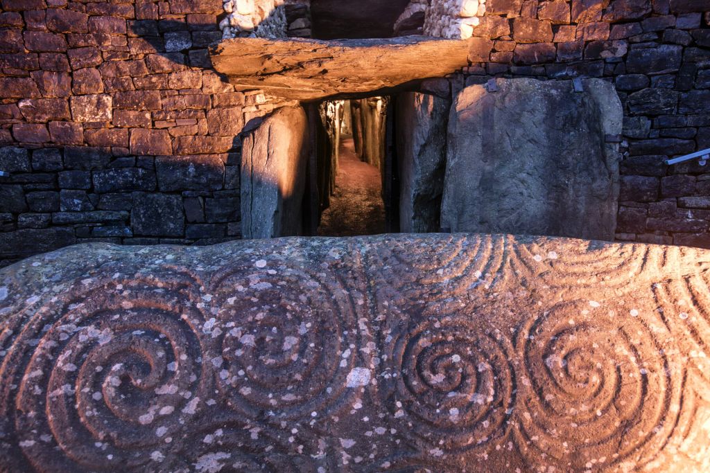 De ingang van een oud stenen graf met een doorgang doet denken aan een rondreis Ierland. Op de voorgrond staat een grote stenen plaat met ingewikkeld gesneden spiraalpatronen. Warm licht verlicht de muren en benadrukt de texturen en houtsnijwerken tegen het schaduwrijke interieur van het graf. | Echt Ierland Vakanties