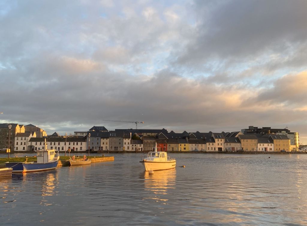 Een serene waterkant bij zonsondergang met kleurrijke rijtjeshuizen langs de kust, weerspiegeld in het kalme water. Een kleine witte boot drijft op de voorgrond en de lucht is gedeeltelijk bewolkt met een warme gouden gloed. In de verte hint een bouwkraan naar nieuwe avonturen op uw reisen Irland. | Echt Ierland Vakanties