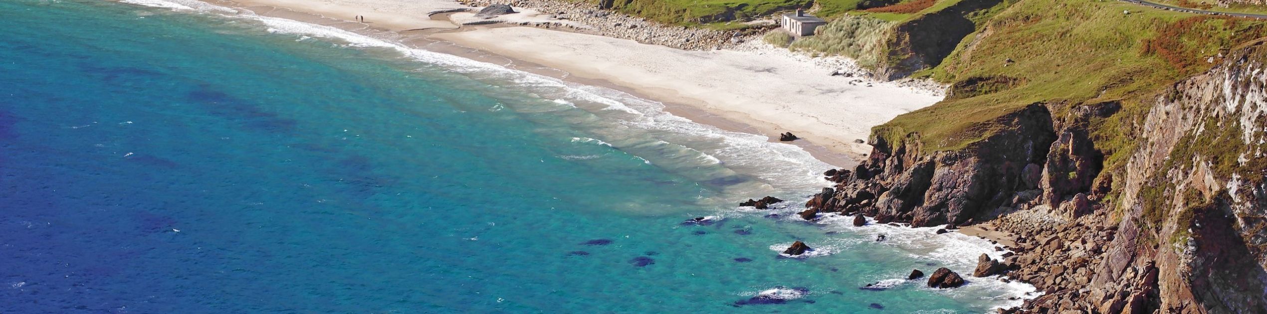 Luchtfoto van een kustlandschap in Ierland met een wit zandstrand omgeven door turquoise wateren en rotsachtige kliffen. De groene, met gras begroeide heuvels rijzen steil op achter het strand, met een kronkelende weg en verspreide gebouwen genesteld in de heuvel, onder een heldere, zonnige hemel - een perfecte plek voor elke vakantie in Ierland. | Echt Ierland Vakanties