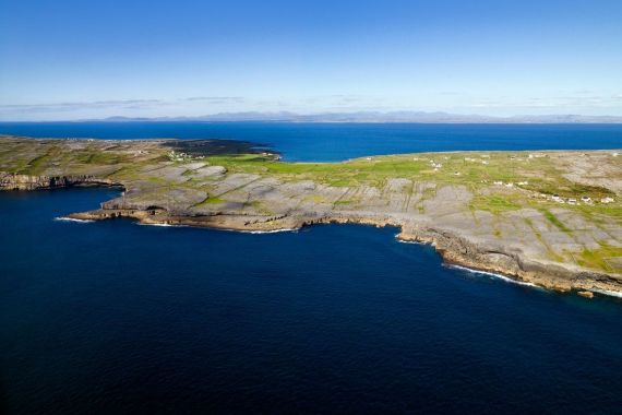 Luchtfoto van een rotsachtige kustlijn in Ierland, met schaars gras en een diepblauwe zee onder een heldere hemel. Het land steekt uit in de oceaan en vormt kliffen, met verspreide kleine witte structuren die deze schilderachtige rondreis Ierland bezaaien. In de verte strekt een kalme oceaan zich uit naar een vage horizon met schaarse wolken. | Echt Ierland Vakanties