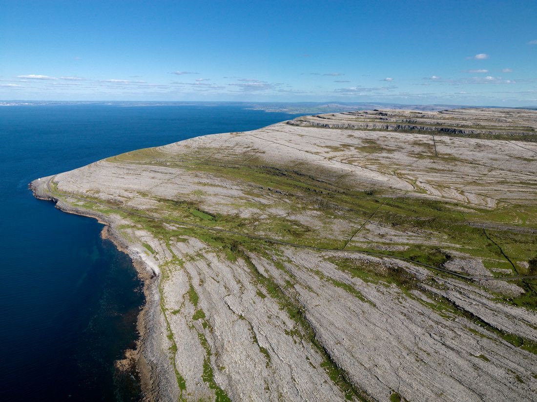 Luchtfoto van een rotsachtig kustlandschap met uitgestrekte kalksteenterrassen die zich uitstrekken tot aan de oceaan, perfect voor een urlaub Ierland. Het terrein wordt afgewisseld met stukken groene vegetatie onder een helderblauwe lucht, die de verre horizon ontmoeten waar water en lucht samenkomen. | Echt Ierland Vakanties
