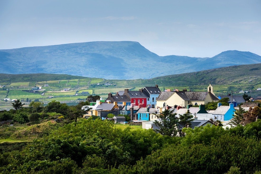 Ervaar de charme van een schilderachtig dorpje in Ierland met kleurrijke huizen in rood, blauw en geel. Weelderig groen omringt dit pittoreske tafereel van glooiende heuvels en bergen onder een heldere hemel. Perfect voor een onvergetelijke rondreis Ierland. | Echt Ierland Vakanties