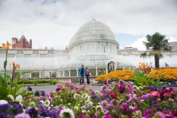 Een stel staat voor een grote, sierlijke glazen serre, die doet denken aan de charmante bouwwerken die je op vakantie in Ierland vindt. Omringd door levendige tuinen met paarse, witte en oranje bloemen en een hoge palmboom aan de rechterkant, genieten ze van een tafereel dat wordt gesierd door een zacht bewolkte lucht. | Echt Ierland Vakanties