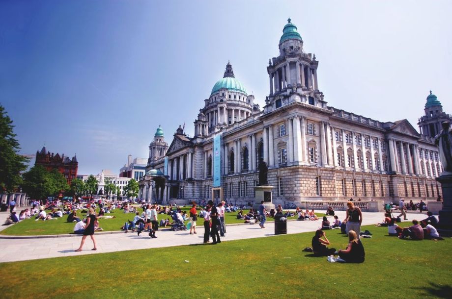 Mensen ontspannen op het gras voor een groot historisch gebouw met een groene koperen koepel, die doet denken aan die van Rundreise Irland. Het tafereel is zonnig met een helderblauwe lucht en de achtergrond bestaat uit bomen en een ander sierlijk gebouw met puntige daken. | Echt Ierland Vakanties