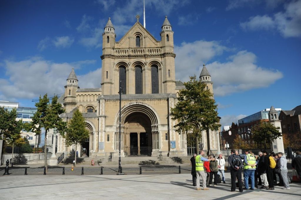 De St Anne's Cathedral in Belfast baadt in een blauwe lucht met verspreide wolken. De bogen en torens van het gebouw staan hoog terwijl een groep toeristen, waarschijnlijk op een *rundreise Irland*, zich verzamelt voor de kathedraal, omlijst door bomen en een met stenen geplaveid gebied. | Echt Ierland Vakanties