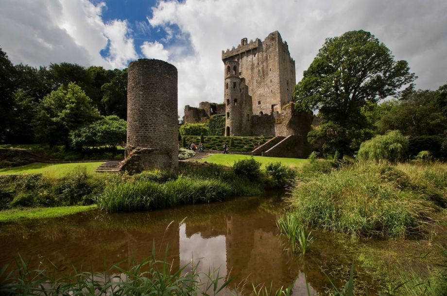 De stenen toren van Blarney Castle rijst op te midden van weelderig groen onder een bewolkte lucht. Op de voorgrond weerspiegelen een ronde toren en een kalme vijver met grasachtige oevers het tafereel, perfect voor degenen die op een rundreise Ierland zijn. Bezoekers slenteren over het pad, omringd door bomen en verzorgde gazons. | Echt Ierland Vakanties