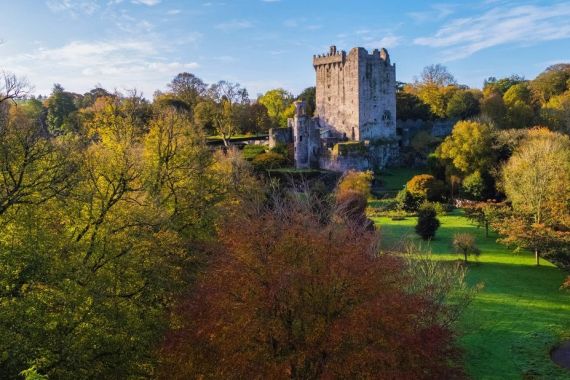 Luchtfoto van een groot, oud stenen kasteel in Ierland, omgeven door weelderige groene velden en dichte, kleurrijke bomen in de vroege herfst. De lucht is helder met een paar dunne wolken. Dit prachtige landschap is een perfect hoogtepunt voor elke rondreis Ierland met zijn levendige tinten groen, oranje en geel gebladerte. | Echt Ierland Vakanties