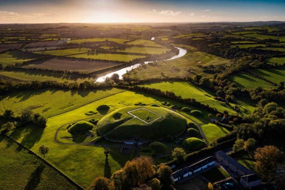Luchtfoto van een weelderig groen landschap in Ierland met een oude heuvel. Een rechthoekige structuur staat erbovenop, omringd door kleinere aarden wallen. Een rivier slingert door de velden en bomen onder een heldere zonsondergang. Nabijgelegen gebouwen geven een hint van de charme die u te wachten staat tijdens uw rondreis Ierland. | Echt Ierland Vakanties