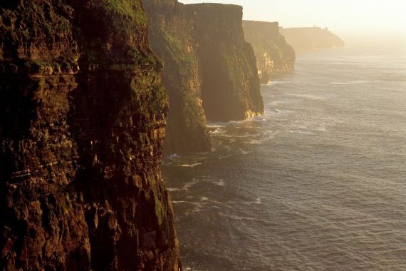 Majestueuze kliffen kijken uit over een uitgestrekte oceaan onder een helderblauwe lucht, die u uitnodigen om ze te verkennen tijdens uw rondreis Ierland. Deze ruige, groene reuzen strekken zich uit tot aan de horizon terwijl zachte golven eronder beuken, het zonlicht werpt een warme gloed. Kleine silhouetten stippelen de kliftoppen, wat wijst op de aantrekkingskracht van reizen Ierland. | Echt Ierland Vakanties