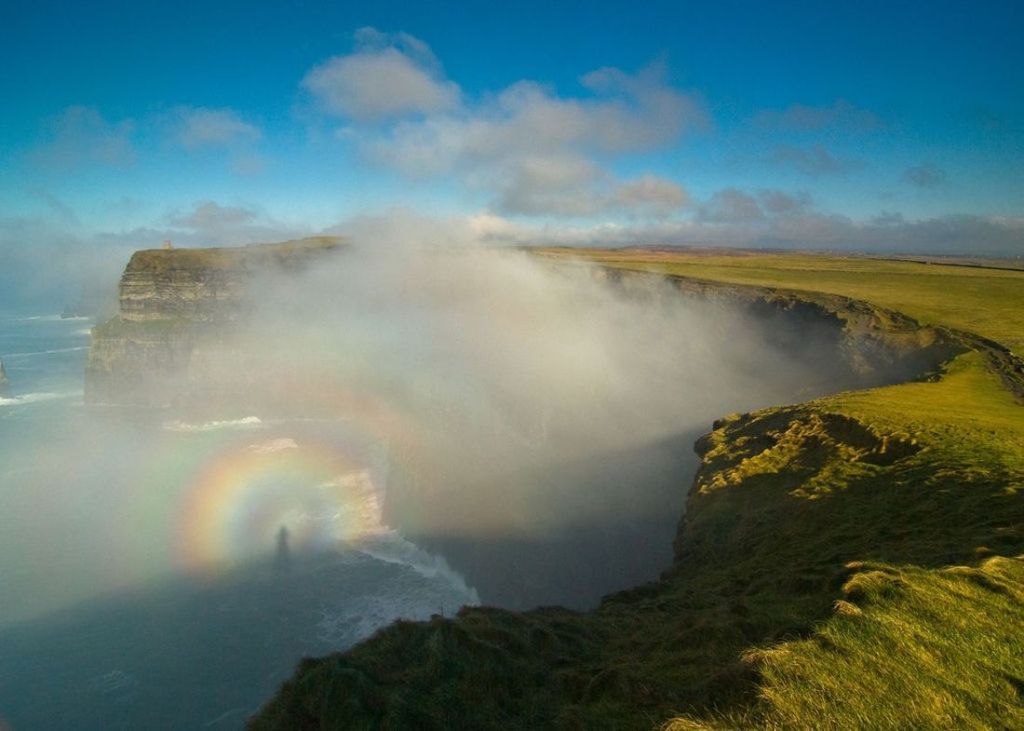 Een mistig kustlandschap in Ierland toont steile kliffen die gedeeltelijk in mist zijn gehuld. Een vage regenboog verschijnt in de mist en werpt een schaduwachtige figuur, mogelijk vanwege het mistboogeffect. De met gras begroeide kliftoppen strekken zich uit tot in de verte onder een blauwe lucht met verspreide wolken - een betoverend tafereel voor vakantie Ierland. | Echt Ierland Vakanties
