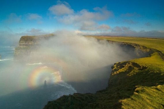 Een mistig kustlandschap in Ierland toont steile kliffen die gedeeltelijk in mist zijn gehuld. Een vage regenboog verschijnt in de mist en werpt een schaduwachtige figuur, mogelijk vanwege het mistboogeffect. De met gras begroeide kliftoppen strekken zich uit tot in de verte onder een blauwe lucht met verspreide wolken - een betoverend tafereel voor vakantie Ierland. | Echt Ierland Vakanties