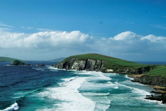 Ervaar het schilderachtige uitzicht op de ruige kustlijn van Ierland, waar turquoise golven tegen de kliffen slaan. Het grasachtige terrein bovenop de kliffen contrasteert met de diepblauwe oceaan. In de verte ontmoeten glooiende heuvels de horizon onder een gedeeltelijk bewolkte lucht, perfect voor uw volgende reisen Irland-avontuur. | Echt Ierland Vakanties