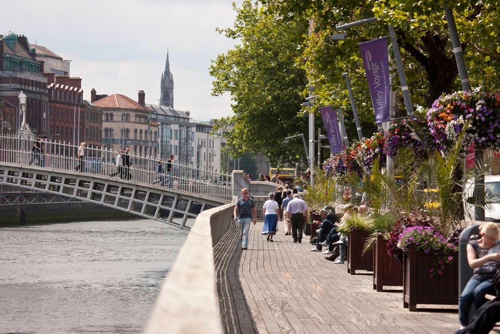 Een levendig riviergezicht met mensen die slenteren en zitten langs een promenade met kleurrijke bloempotten roept de charme van Rundreise Irland op. Op de achtergrond overspant een historische, sierlijke metalen brug de rivier. De lucht is gedeeltelijk bewolkt en weelderige bomen bieden schaduw langs de loopbrug. | Echt Ierland Vakanties