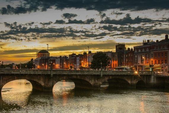 Een schilderachtig uitzicht op een stadsbrug over een kalme rivier bij zonsondergang, wat doet denken aan de pittoreske landschappen die je vaak ziet tijdens een vakantie in Ierland. De lucht heeft dramatische wolken en warme tinten oranje en geel. Historische gebouwen met verlichte ramen langs de oever van de rivier, weerspiegeld in het water onder de verlichte bogen. | Echt Ierland Vakanties