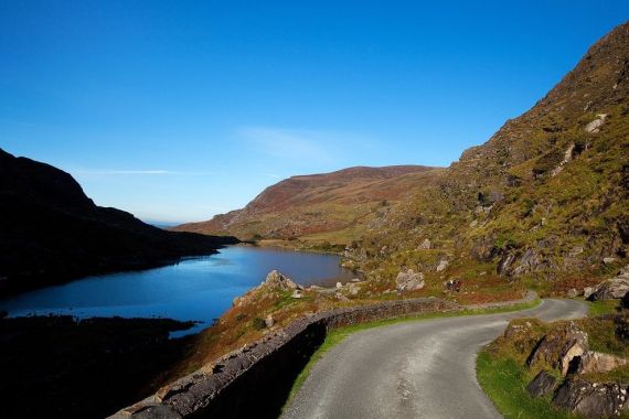 Een kronkelende bergweg in Ierland slingert langs een kalm blauw meer onder een helderblauwe lucht. Het landschap, perfect voor een Ierland vakantieavontuur, bestaat uit rotsachtige heuvels met schaarse vegetatie en stukken groen en bruin. Verre bergtoppen koesteren zich in het zonlicht aan de horizon. | Echt Ierland Vakanties