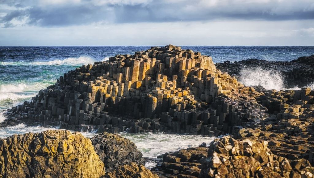 De afbeelding legt de Giant's Causeway vast, een adembenemend hoogtepunt van elke rondreis Ierland. Talloze hexagonale basaltkolommen rijzen majestueus op uit de woelige, diepblauwe zee terwijl golven er tegenaan beuken onder een gedeeltelijk bewolkte lucht. | Echt Ierland Vakanties