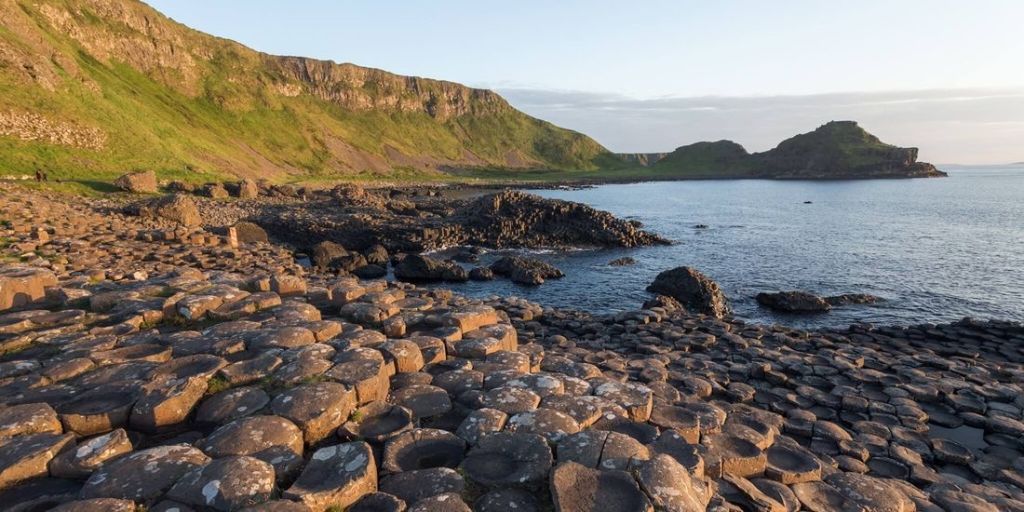 De afbeelding toont de Giant's Causeway in Noord-Ierland, een must-visit op elke rundreise in Ierland. In elkaar grijpende basaltkolommen strekken zich uit tot aan de zee, badend in warm zonlicht. Groene kliffen rijzen majestueus op tegen een kalme oceaan, verdwijnend in de horizon onder een gedeeltelijk bewolkte lucht. | Echt Ierland Vakanties