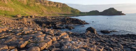 De afbeelding toont de Giant's Causeway in Noord-Ierland, een must-visit op elke rundreise in Ierland. In elkaar grijpende basaltkolommen strekken zich uit tot aan de zee, badend in warm zonlicht. Groene kliffen rijzen majestueus op tegen een kalme oceaan, verdwijnend in de horizon onder een gedeeltelijk bewolkte lucht. | Echt Ierland Vakanties