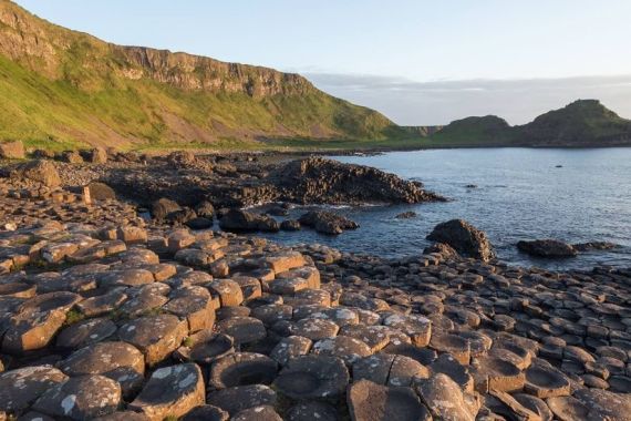 De afbeelding toont de Giant's Causeway in Noord-Ierland, een must-visit op elke rundreise in Ierland. In elkaar grijpende basaltkolommen strekken zich uit tot aan de zee, badend in warm zonlicht. Groene kliffen rijzen majestueus op tegen een kalme oceaan, verdwijnend in de horizon onder een gedeeltelijk bewolkte lucht. | Echt Ierland Vakanties