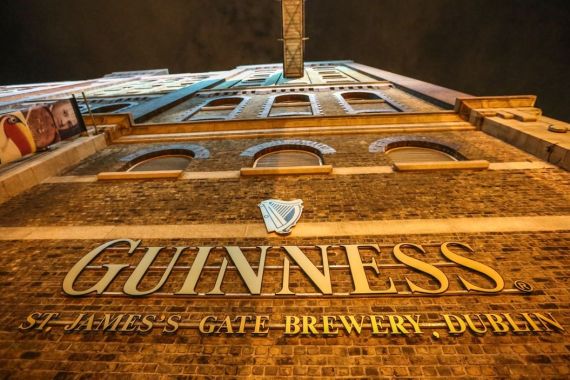 Laag standpunt van het Guinness Storehouse in Dublin bij nacht, met de bakstenen gevel. Het gebouw heeft een groot, verlicht Guinness harp-logo met de tekst "Guinness St. James's Gate Brewery, Dublin" in gouden letters. Een must-see op elke rundreise in Ierland onder bewolkte, donkere luchten. | Echt Ierland Vakanties