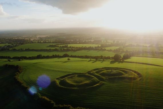 Luchtfoto van een weelderig groen landschap in Ierland bij zonsondergang, met twee cirkelvormige aarden wallen te midden van velden. Schaduwen van de laagstaande zon strekken zich uit over het terrein, met verspreide bomen die charme toevoegen. Een zonnevlam verlicht het tafereel en versterkt de serene sfeer die perfect is voor een vakantie in Ierland. | Echt Ierland Vakanties