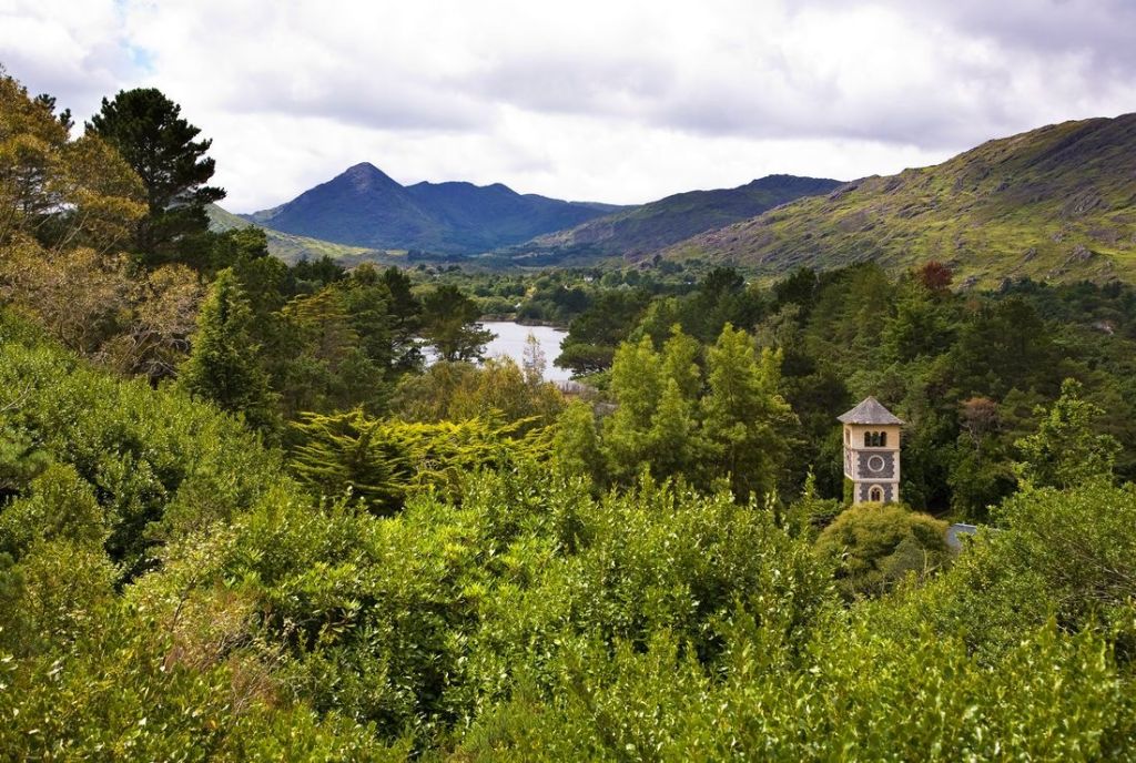 Een weelderig landschap met een beboste vallei en een rivier op de middengrond roept de serene schoonheid van reizen Ierland op. Een hoge, smalle toren met een puntig dak steekt door de bomen heen. Heuvels en bergen rijzen op de achtergrond op onder een gedeeltelijk bewolkte lucht, waardoor diepte en rust ontstaat. | Echt Ierland Vakanties