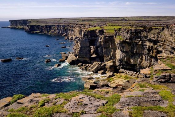 De ruige kustlijn van een rundreise Ierland-avontuur onthult rotsachtige kliffen die baden in zonlicht. De diepblauwe oceaan beukt tegen de oude klifbasis, terwijl stukken groen gras hun toppen bekronen. Dunne wolken stippelen de heldere lucht en versterken dit schilderachtige uitzicht. | Echt Ierland Vakanties