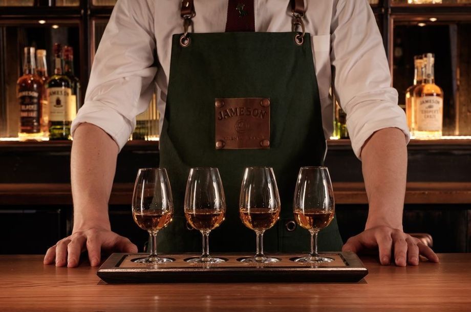 Een persoon met een groene schort staat achter een houten bar, wat de charme van een vakantie in Ierland oproept. Vier glazen whisky staan op een klein houten dienblad voor hen, terwijl flessen sterke drank de planken vullen in de zacht verlichte achtergrond. | Echt Ierland Vakanties