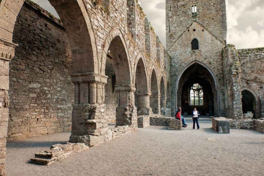 Een historische ruïne van een stenen abdij met grote bogen en een hoge toren, perfect voor mensen die een rondreis door Ierland maken. Twee mensen staan in het midden, een in een rood shirt en de ander in het wit, en verkennen deze betoverende plek. De abdij wordt omlijst door een bewolkte lucht en grind bedekt de grond. | Echt Ierland Vakanties