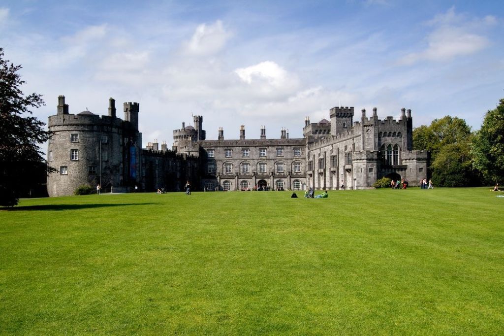 Een historisch stenen kasteel met meerdere torentjes en kantelen, afgezet tegen een bewolkte blauwe lucht. Omringd door een verzorgd gazon met verspreide bezoekers, is dit pittoreske tafereel ideaal voor degenen die een rondreis Ierland maken of genieten van een vakantie in Ierland te midden van weelderig groen. | Echt Ierland Vakanties