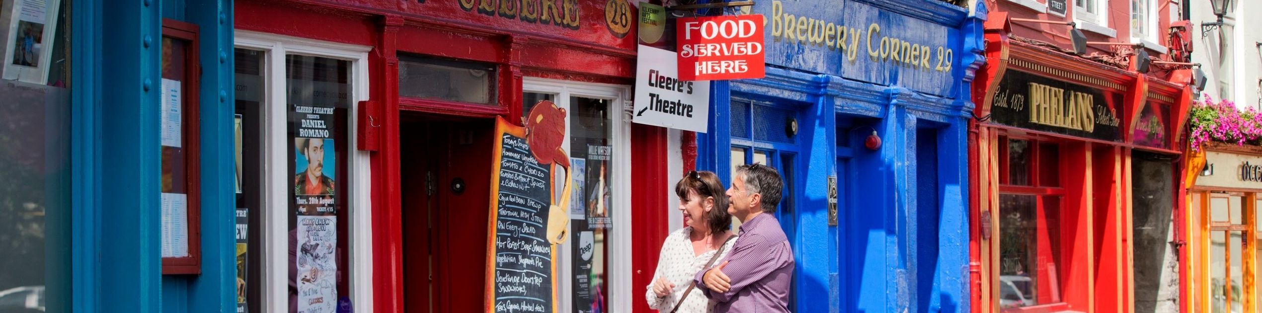 Een stel staat buiten een felrode winkel met het bord "John Culbert's", genietend van de charme van hun rundreise Ierland. Ze lezen een schoolbordmenu terwijl blauwe en gele winkelpuien de straat opfleuren. Hangende bloemenmanden zorgen voor groen, terwijl een bord hen uitnodigt om te verkennen: "Hier wordt eten geserveerd. | Echt Ierland Vakanties