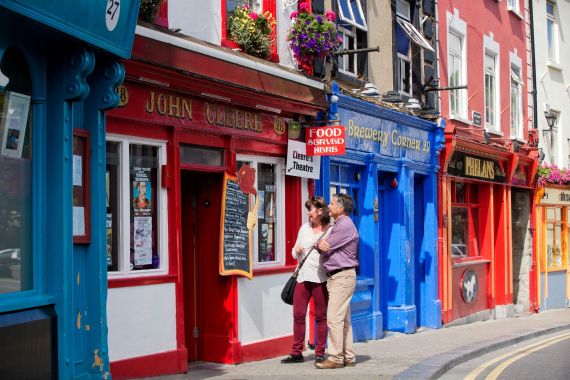 Een stel staat buiten een felrode winkel met het bord "John Culbert's", genietend van de charme van hun rundreise Ierland. Ze lezen een schoolbordmenu terwijl blauwe en gele winkelpuien de straat opfleuren. Hangende bloemenmanden zorgen voor groen, terwijl een bord hen uitnodigt om te verkennen: "Hier wordt eten geserveerd. | Echt Ierland Vakanties