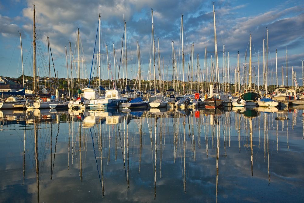 Een jachthaven vol met talloze zeilboten en jachten weerspiegelt op kalm water onder een gedeeltelijk bewolkte lucht, wat doet denken aan serene Ierse landschappen. Masten creëren verticale lijnen tegen het blauw, terwijl de kustlijn op de achtergrond herinneringen oproept aan een vredige vakantie in Ierland. | Echt Ierland Vakanties