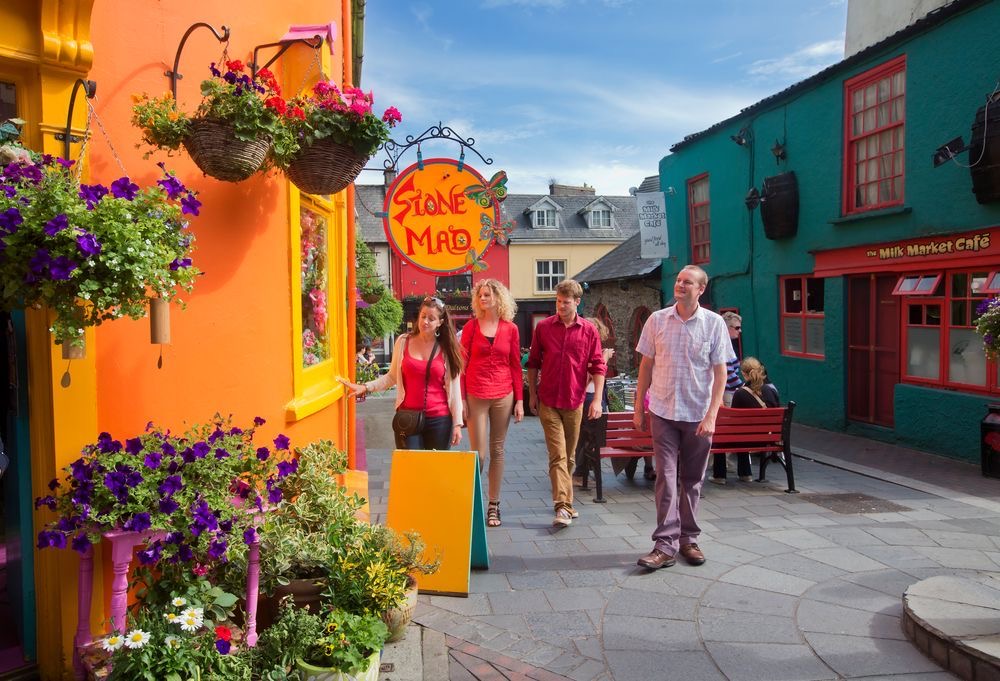 Een kleurrijk straatbeeld in Ierland toont een groep van vier mensen die langs levendige gebouwen lopen. Een oranje gebouw met hangende bloemenmanden en een bord met de tekst "Stone Mad" staat links. Rechts omlijsten turquoise en rode gebouwen een rode bank en café, perfect voor een idyllische Urlaub Irland-ervaring onder een heldere hemel. | Echt Ierland Vakanties