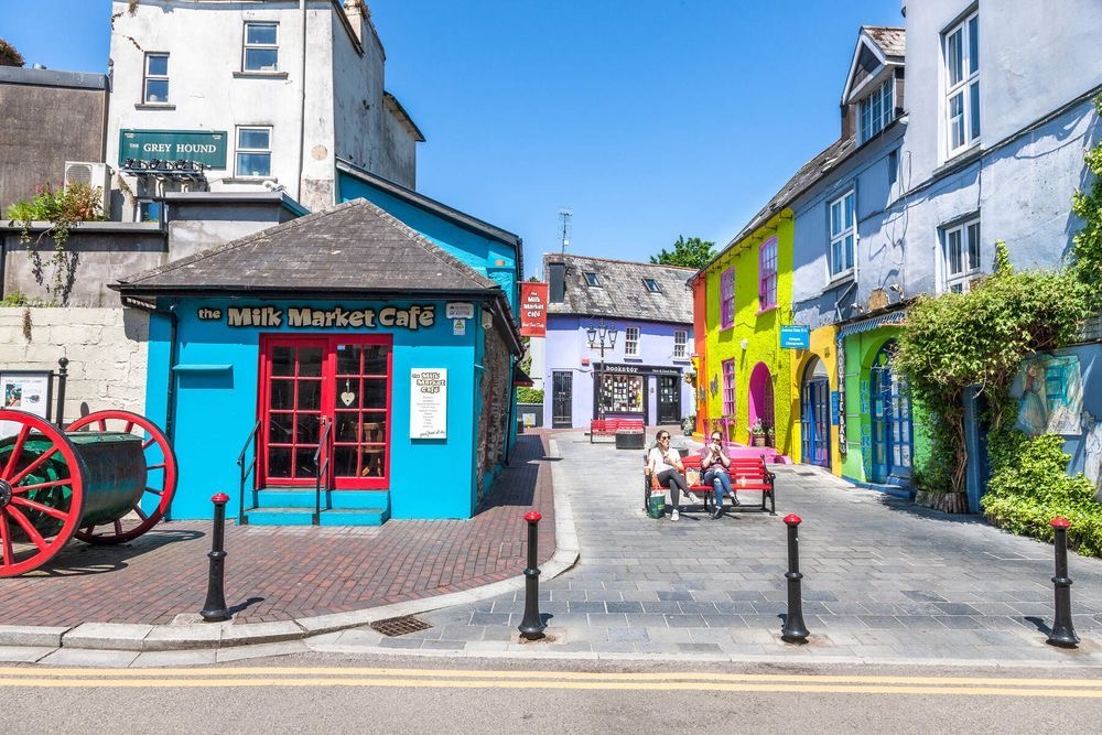 Een levendig straatbeeld vangt de essentie van *vakantie Ierland*, met kleurrijke gebouwen onder een helderblauwe lucht. Het Milk Market Café staat felblauw aan de linkerkant, terwijl gele, paarse en blauwe gevels zich uitstrekken langs de rechterkant. Bezoekers zitten aan buitentafels naast een rustiek rood karrewiel. | Echt Ierland Vakanties