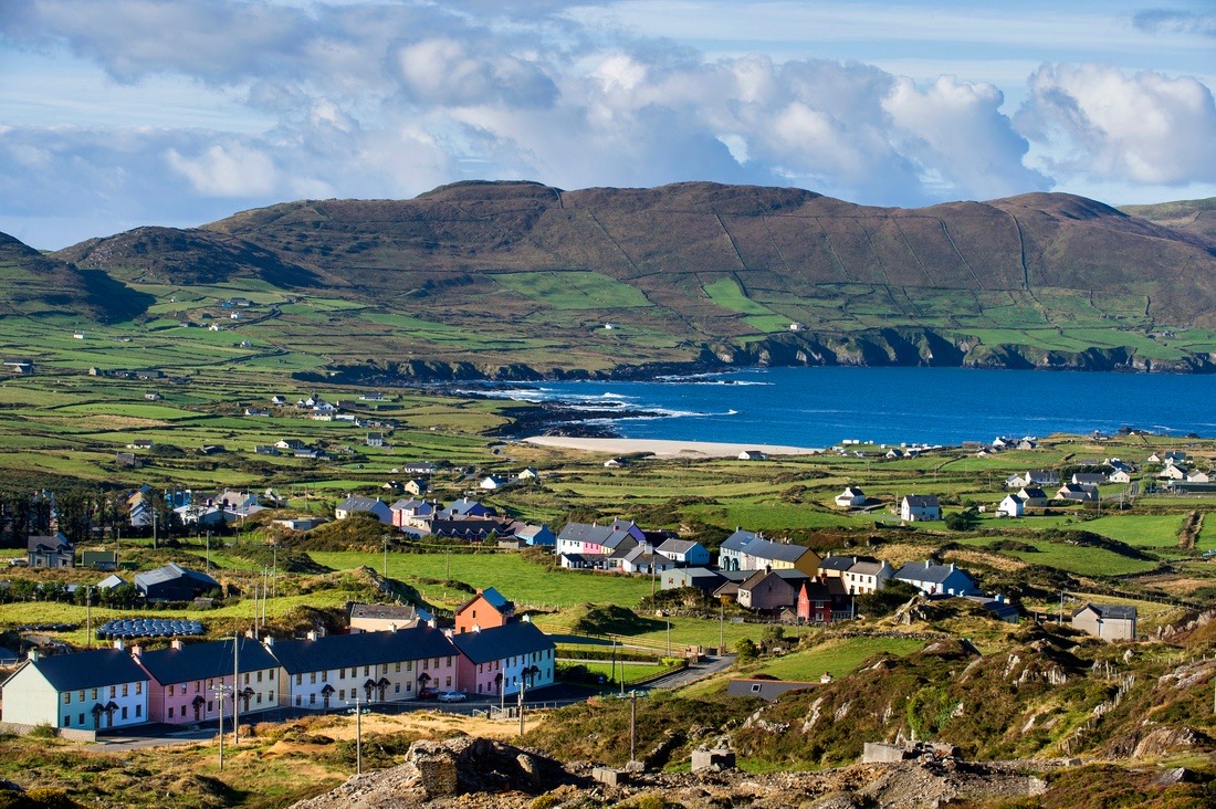 Een schilderachtig uitzicht op een kustdorp in Ierland met kleurrijke huizen verspreid over weelderige groene velden. Het landschap omvat verre heuvels, een kronkelende kustlijn en de diepblauwe zee onder een lucht met verspreide wolken, perfect voor een onvergetelijke "urlaub Irland". | Echt Ierland Vakanties