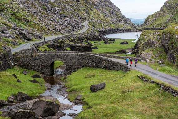 Een schilderachtig landschap dat typerend is voor een rondreis Ierland, met een smalle, kronkelende weg geflankeerd door rotsachtige heuvels en grasachtig terrein. Een stenen brug overspant een kleine beek. Drie wandelaars in kleurrijke outdoorkleding lopen over het pad. Een kalm meer is zichtbaar aan de rechterkant, onder een bewolkte lucht. | Echt Ierland Vakanties