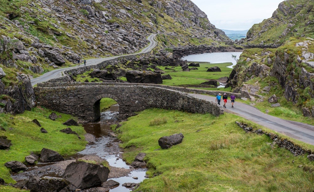 Een schilderachtig landschap dat typerend is voor een rondreis Ierland, met een smalle, kronkelende weg geflankeerd door rotsachtige heuvels en grasachtig terrein. Een stenen brug overspant een kleine beek. Drie wandelaars in kleurrijke outdoorkleding lopen over het pad. Een kalm meer is zichtbaar aan de rechterkant, onder een bewolkte lucht. | Echt Ierland Vakanties