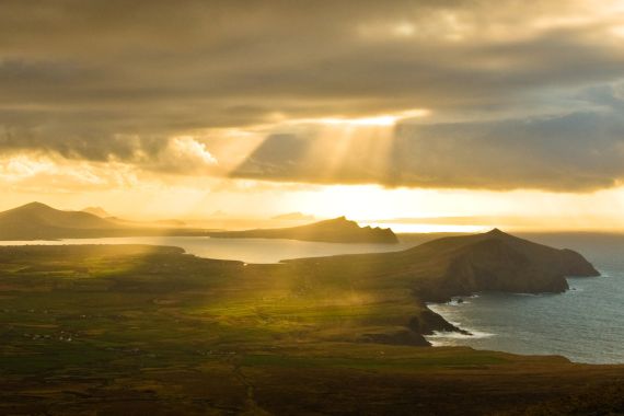 Zonlicht breekt door dramatische wolken en werpt gouden stralen over een kustlandschap met weelderige groene velden en ruige heuvels. Tijdens een rondreis Ierland weerkaatst de oceaan het licht terwijl verre eilanden zich aftekenen tegen de horizon, wat een rustig en pittoresk tafereel creëert. | Echt Ierland Vakanties