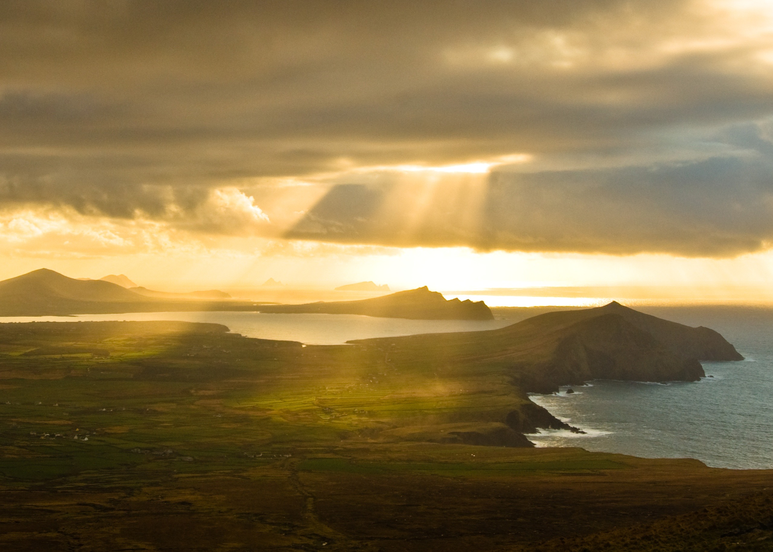 Zonlicht breekt door dramatische wolken en werpt gouden stralen over een kustlandschap met weelderige groene velden en ruige heuvels. Tijdens een rondreis Ierland weerkaatst de oceaan het licht terwijl verre eilanden zich aftekenen tegen de horizon, wat een rustig en pittoresk tafereel creëert. | Echt Ierland Vakanties