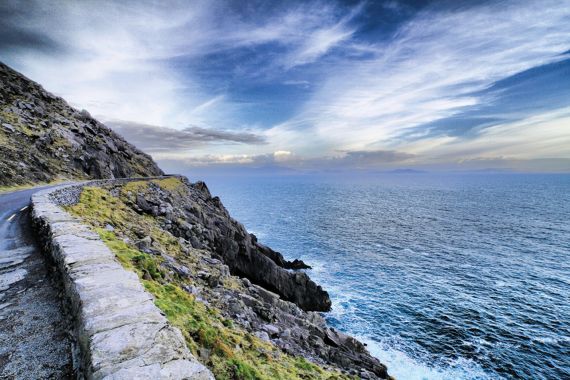 Een kronkelende kustweg in Ierland loopt langs een steile rotsachtige klif aan de linkerkant, begrensd door een lage stenen muur. Aan de rechterkant strekt een uitgestrekte kalme oceaan zich uit tot aan de horizon onder een dramatische lucht met strepen wolken in blauwe en witte tinten - perfect voor een onvergetelijke reis door Ierland. | Echt Ierland Vakanties