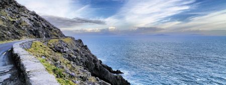 Een kustweg in Ierland slingert langs een steile, rotsachtige klif met uitzicht op een uitgestrekte zee. De lucht is gedeeltelijk bewolkt met verschillende tinten blauw en wit. Golven beuken tegen de voet van de klif, wat een gevoel van serene isolatie en natuurlijke schoonheid biedt - ideaal voor een urlaub Ierland-reis. | Echt Ierland Vakanties