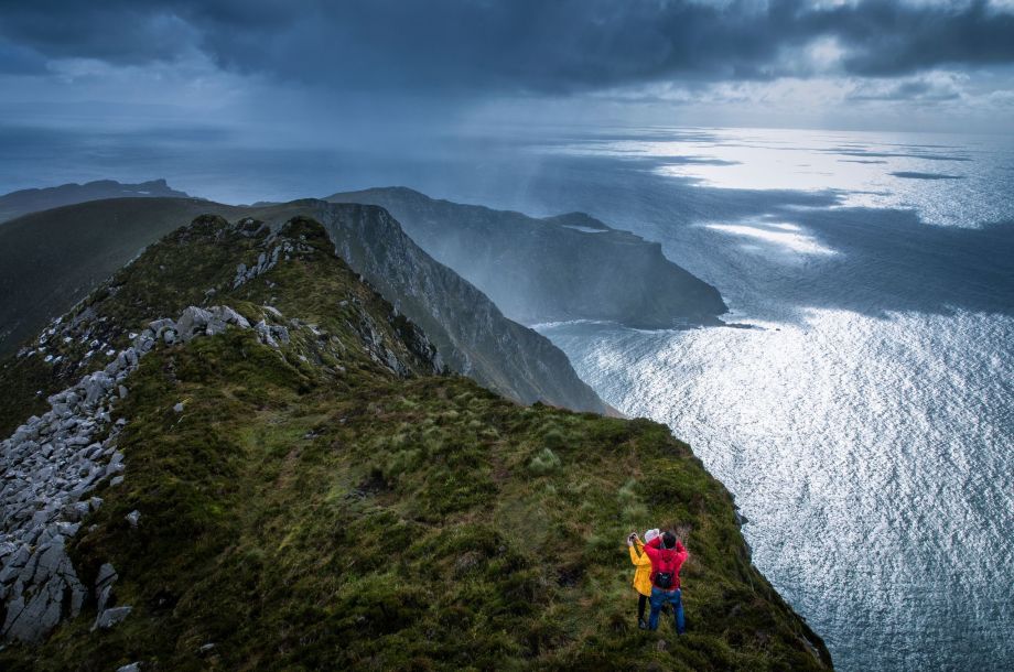 Twee mensen in felle jassen staan op een met gras begroeide klifrand, wat de aantrekkingskracht van Ierland vakanties illustreert. De oceaan glinstert onder een bewolkte, dramatische lucht, met regen zichtbaar in de verte. Bergen strekken zich uit tot in het water, en creëren een ruige en serene kustlijn die perfect is voor een vakantie in Ierland. | Echt Ierland Vakanties