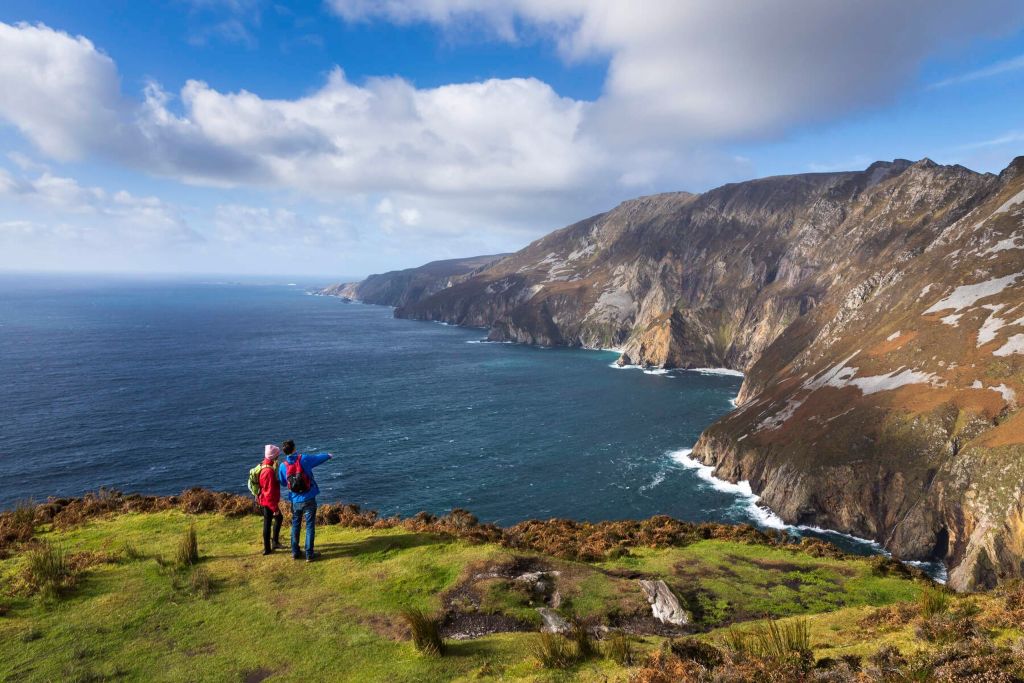 Twee mensen staan op een grasachtige klif in Ierland, één wijst naar de rotsachtige kustlijn. Het tafereel kijkt uit over een uitgestrekte oceaan waar golven tegen ruige kliffen beuken onder een gedeeltelijk bewolkte lucht. Ideaal voor degenen die op zoek zijn naar een rundreise Ierland, dit landschap heeft weelderige stukken oranje en bruin gebladerte. | Echt Ierland Vakanties