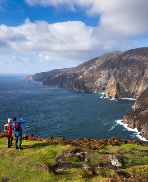 Twee mensen staan op een grasachtige klif in Ierland, één wijst naar de rotsachtige kustlijn. Het tafereel kijkt uit over een uitgestrekte oceaan waar golven tegen ruige kliffen beuken onder een gedeeltelijk bewolkte lucht. Ideaal voor degenen die op zoek zijn naar een rundreise Ierland, dit landschap heeft weelderige stukken oranje en bruin gebladerte. | Echt Ierland Vakanties