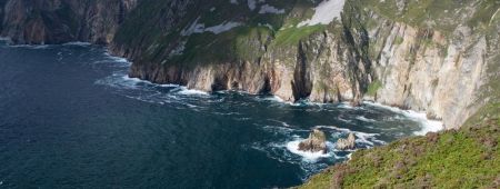 Een schilderachtig uitzicht op torenhoge zeekliffen die de oceaan ontmoeten, een typisch gezicht tijdens een rondreis Ierland. De kliffen, bedekt met groen, rijzen steil op boven donkerblauw water waar golven tegen rotsen beuken. De voorgrond pronkt met heideachtige vegetatie onder een gedeeltelijk bewolkte lucht, wat de essentie van vakantie Ierland vastlegt. | Echt Ierland Vakanties